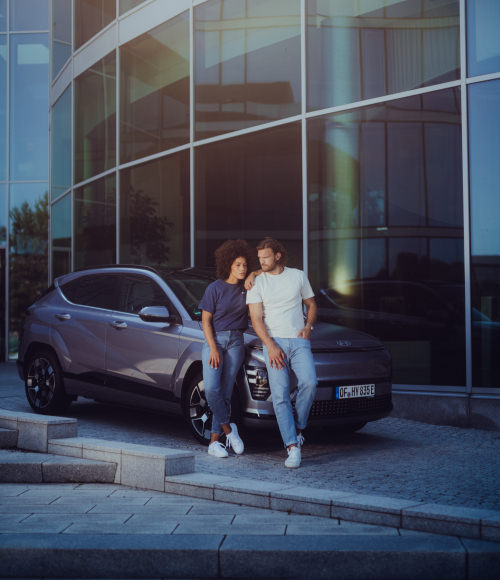 A woman and a man, standing next to a gray Hyundai SUV in front of a modern glass building, with the text 'Hyundai Collection' at the bottom.