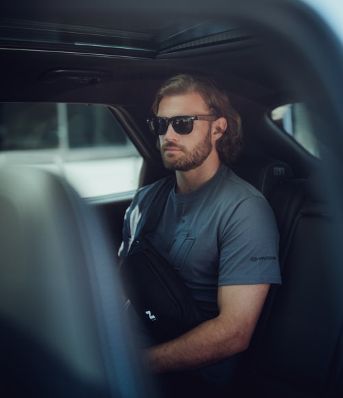 A man in sunglasses and a grey shirt sits in a car. 'New' is in the corner.