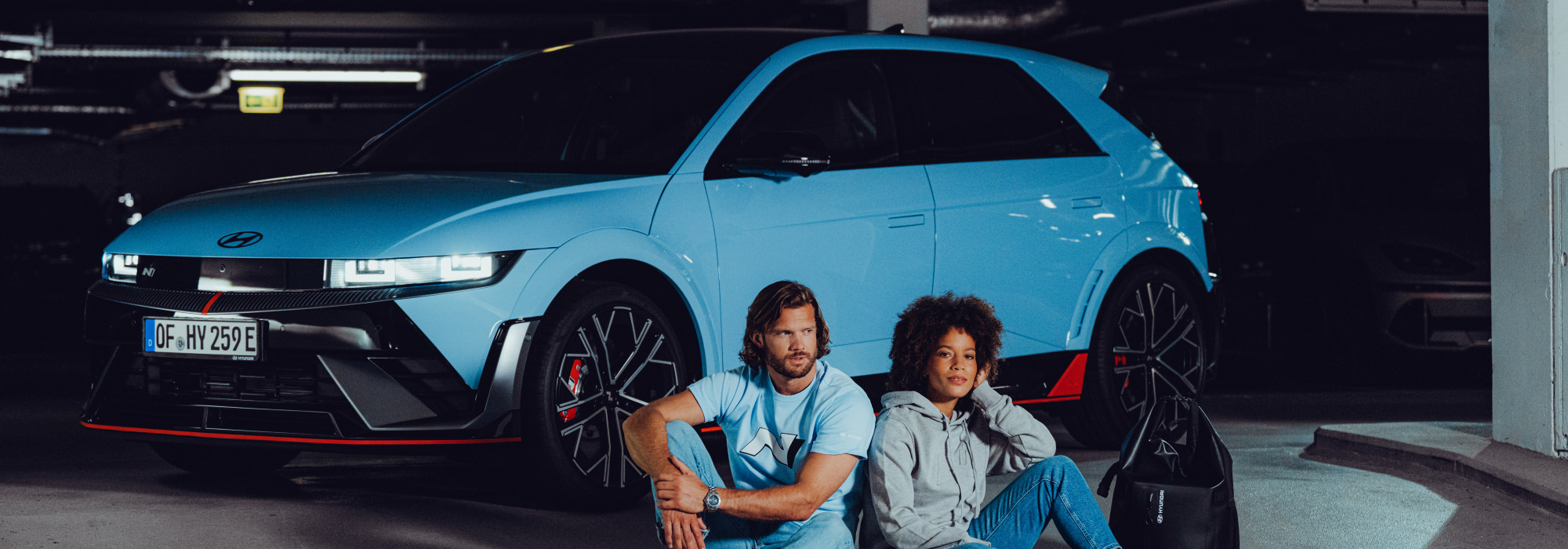 Two people sitting in front of a blue Hyundai N car in a dark parking garage.