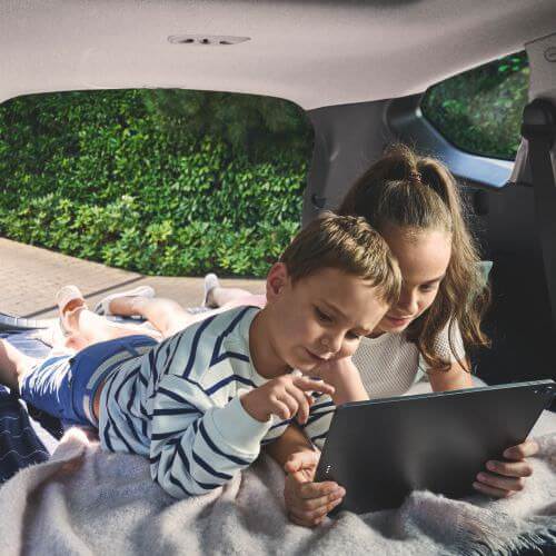 Two children are lying in the back of a car, closely looking at a tablet together, with greenery visible outside the window.